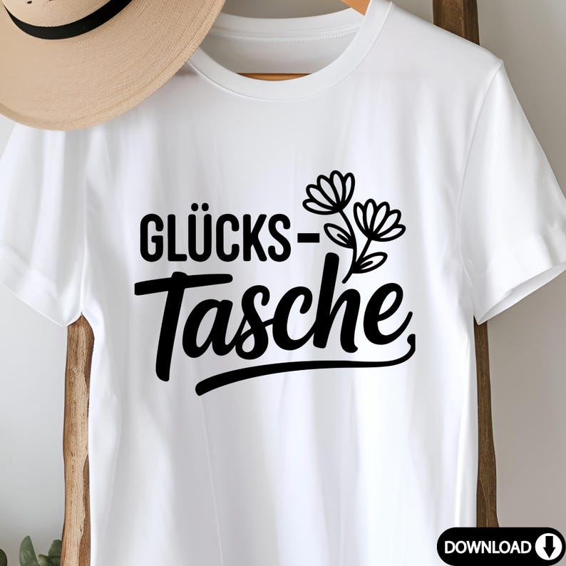 May include: White t-shirt featuring the German text "GL&Uuml;CKS-Tasche" in black, with a floral illustration. The shirt is made of a soft material and is suitable for casual wear. A straw hat is partially visible in the background.