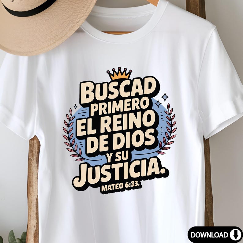 May include: White t-shirt with a colorful graphic design. The design features the text "Buscad Primero El Reino De Dios Y Su Justicia" in a stylized font, with a crown and laurel wreath. The text "Mateo 6:33" is also included.