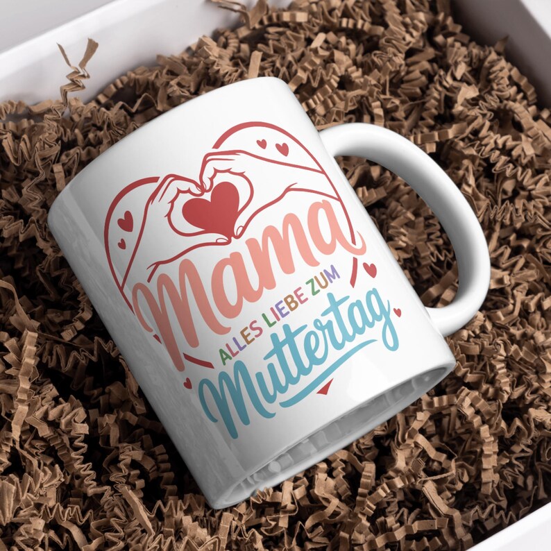May include: White ceramic mug with a heart-shaped design made with hands and a red heart. The word "Mama" is written in a pink script, with the text "Alles Liebe zum Muttertag" in blue and red.