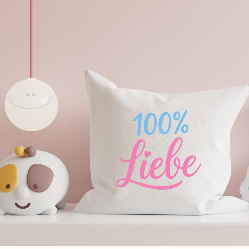 May include: White throw pillow with the text "100% Liebe" in blue and pink script. A decorative smiling light fixture hangs above a white shelf. A white and brown toy is on the shelf.