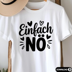 Puede incluir: Camiseta blanca con la frase alemana "Einfach N&ouml;" en negro, decorada con corazones y dise&ntilde;os de rayos de sol. Una camiseta casual de manga corta, perfecta para el uso diario. Un sombrero de paja es parcialmente visible en el fondo.