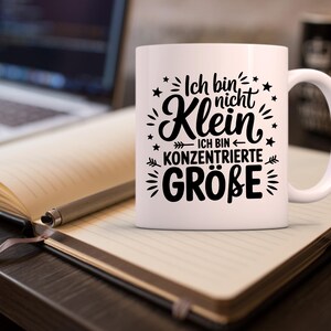 Puede incluir: Taza de cer&aacute;mica blanca con texto negro "Ich bin nicht Klein - Ich bin konzentrierte Gr&ouml;&szlig;e" rodeado de estrellas y elementos decorativos. La taza est&aacute; sobre un cuaderno con un bol&iacute;grafo, con un port&aacute;til al fondo.