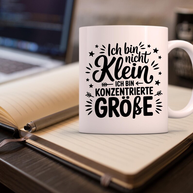 Puede incluir: Taza de cer&aacute;mica blanca con texto negro "Ich bin nicht Klein - Ich bin konzentrierte Gr&ouml;&szlig;e" rodeado de estrellas y elementos decorativos. La taza est&aacute; sobre un cuaderno con un bol&iacute;grafo, con un port&aacute;til al fondo.