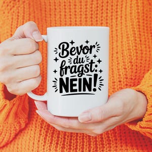 Puede incluir: Taza de cer&aacute;mica blanca con la frase alemana "Bevor du fragst: NEIN!" en escritura negra. La taza tiene un asa grande y la sostiene una persona con un su&eacute;ter naranja. El texto est&aacute; rodeado de estrellas decorativas.