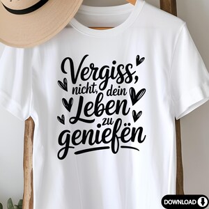 May include: White t-shirt featuring the German phrase "Vergiss nicht, dein Leben zu genie&szlig;en" in black script, meaning "Don't forget to enjoy your life." The design includes heart accents. A straw hat is partially visible in the background.