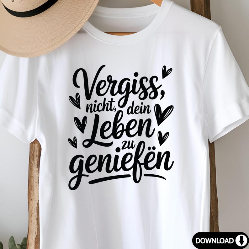 May include: White t-shirt featuring the German phrase "Vergiss nicht, dein Leben zu genie&szlig;en" in black script, meaning "Don't forget to enjoy your life." The design includes heart accents. A straw hat is partially visible in the background.