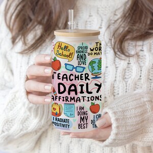 Op de afbeelding: Een witte beker met een bamboe deksel en een rietje. De beker is versierd met kleurrijke illustraties en de tekst "Teacher Daily Affirmations". De illustraties omvatten een wereldbol, een schoolbus, een potlood, een boek en een appel.