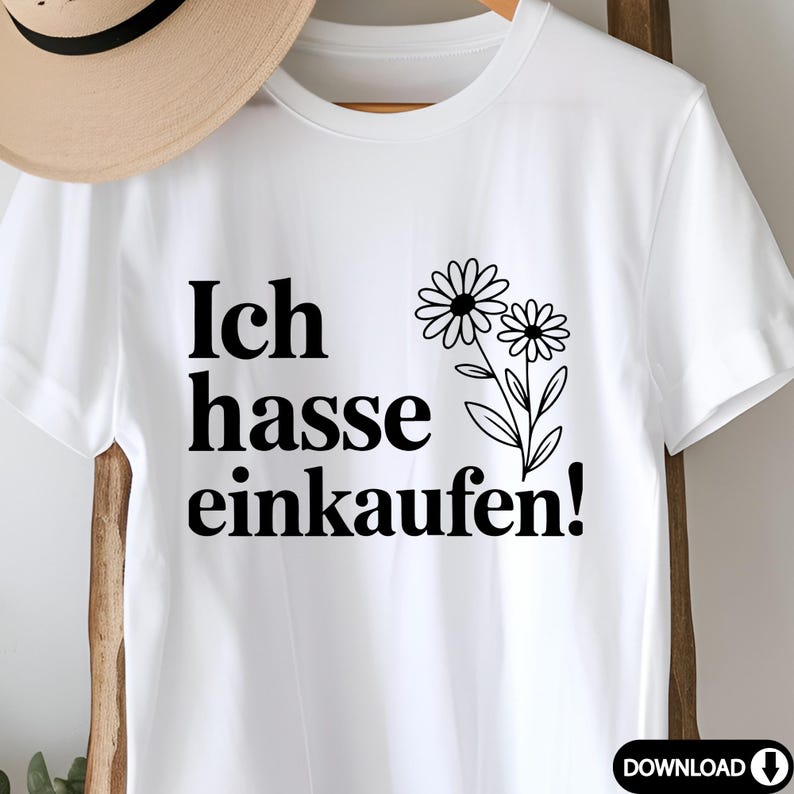 May include: White t-shirt with the German phrase "Ich hasse einkaufen!" in black, meaning "I hate shopping!" accompanied by a black floral illustration. The shirt has short sleeves and a crew neck.