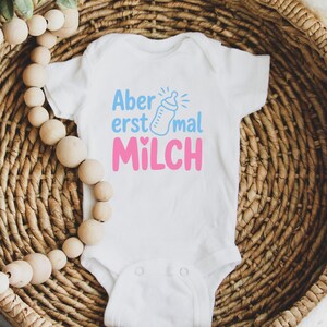May include: White baby onesie with the German text "Aber erst mal MILCH" in blue and pink. The design includes a baby bottle illustration. The onesie is displayed on a woven basket.