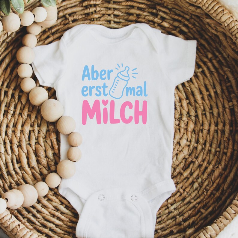 May include: White baby onesie with the German text "Aber erst mal MILCH" in blue and pink. The design includes a baby bottle illustration. The onesie is displayed on a woven basket.