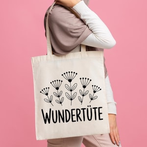 May include: A cream-colored tote bag featuring a black floral design and the word "WUNDERTIITE" in bold, black letters. The bag has two handles and is held against a pink background.