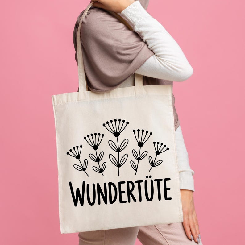 May include: A cream-colored tote bag featuring a black floral design and the word "WUNDERTIITE" in bold, black letters. The bag has two handles and is held against a pink background.