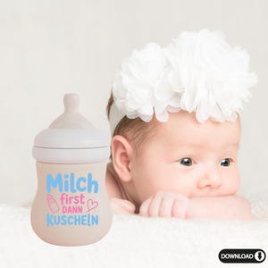 May include: A beige baby bottle with a white top and a light blue and pink design that reads "Milch first dann Kuscheln" with a baby bottle graphic and a heart. A baby wearing a white floral headband is in the background.