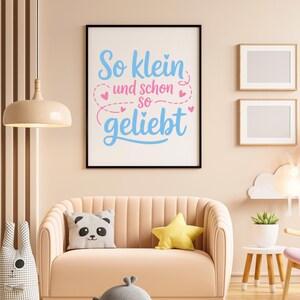 May include: A framed print with the text "So klein und schon so geliebt" in blue and pink. The print hangs above a peach-colored sofa with a panda pillow and a yellow star-shaped pillow. A decorative lamp and two framed pictures are also visible.