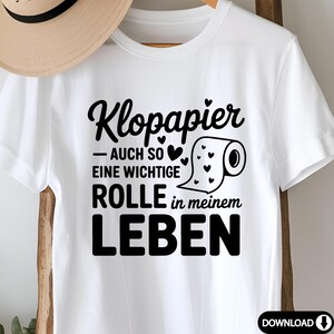 Puede incluir: Camiseta blanca con texto negro "Klopapier - AUCH SO EINE WICHTIGE ROLLE in meinem LEBEN" y un gr&aacute;fico de un rollo de papel higi&eacute;nico con corazones. Un sombrero beige cuelga en el fondo.
