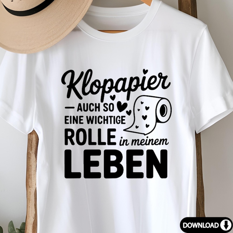 Puede incluir: Camiseta blanca con texto negro "Klopapier - AUCH SO EINE WICHTIGE ROLLE in meinem LEBEN" y un gr&aacute;fico de un rollo de papel higi&eacute;nico con corazones. Un sombrero beige cuelga en el fondo.