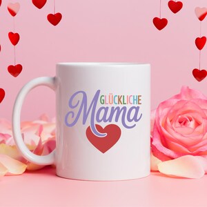 May include: White ceramic mug with the words "Mama" and "Gl&uuml;ckliche" in a script font, with a red heart. The mug is set against a pink background with hanging red heart decorations and a pink rose.