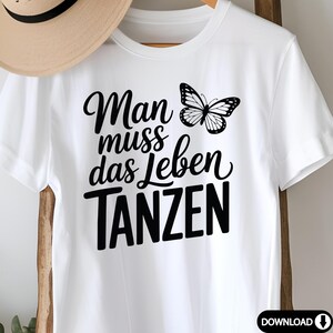 May include: White t-shirt with the German phrase "Man muss das Leben TANZEN" and a black butterfly graphic. The text translates to "One must dance life". A tan hat is partially visible in the background.