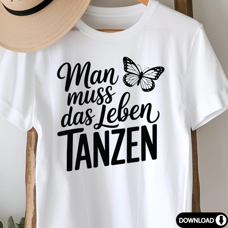 May include: White t-shirt with the German phrase "Man muss das Leben TANZEN" and a black butterfly graphic. The text translates to "One must dance life". A tan hat is partially visible in the background.