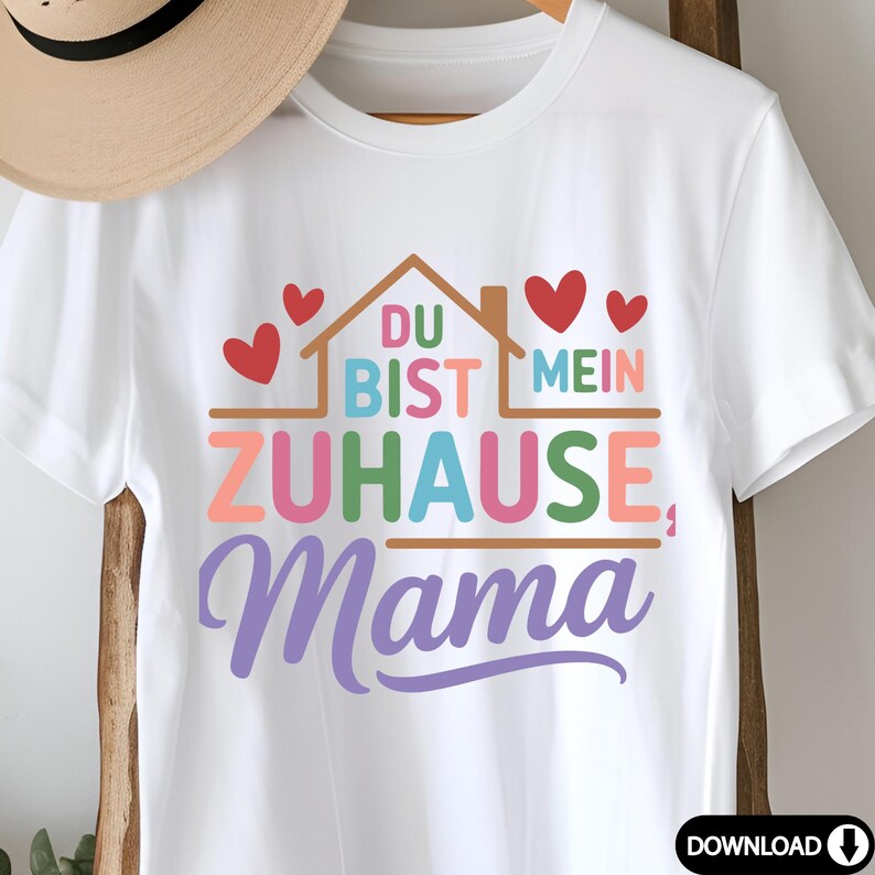 May include: White t-shirt with the German phrase "Du bist mein Zuhause, Mama" in colorful text and a house graphic with red hearts. The design is printed on a classic crew neck tee.