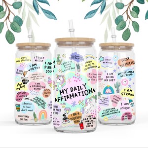 Op de afbeelding: Drie glazen waterflessen met een transparante kleur, bamboe deksels en rietjes. Elke fles heeft kleurrijke stickers met positieve affirmaties en illustraties van eenhoorns, cactussen en regenbogen. De tekst op de stickers bevat zinnen als "Ik ben het waard", "Ik doe mijn best" en "Mijn dagelijkse affirmaties".