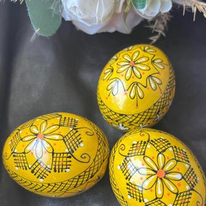 Op de afbeelding: Drie handgeschilderde decoratieve paaseieren. De eieren zijn geel met ingewikkelde zwart-witte bloemen- en geometrische ontwerpen. De ontwerpen omvatten bloemen, stippen en rasterpatronen. De eieren zijn gerangschikt op een donkere ondergrond.