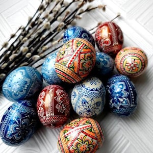 Peut inclure: Une collection d'œufs de Pâques décorés de couleurs vives, dans les tons de bleu, rouge, orange et blanc. Chaque œuf présente des motifs et des dessins complexes. Un bouquet de chatons est en arrière-plan.
