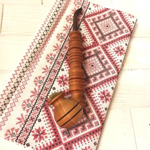 Puede incluir: Una pipa de fumar de madera con un acabado marrón y negro. La pipa tiene una cazoleta redonda y una boquilla larga y curva. La pipa está apoyada sobre una tela blanca con un patrón geométrico rojo y negro.