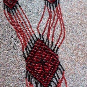 May include: A red and black beaded necklace with a geometric pattern. The necklace has multiple strands and a fringe at the bottom.