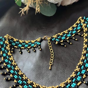 以下が含まれることがあります： 黒、ターコイズ、ゴールドのデザインのビーズネックレス。ネックレスは、ターコイズとゴールドのビーズの繰り返し模様で、黒いビーズが縁取りをしています。留め金とチェーンは金色です。ネックレスは暗い表面に表示されています。