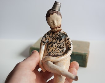 The girl with the thimble to sit on books, shelf shelf. Ceramic figure. Sculpture of woman sitting with crossed legs