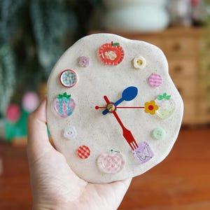 Può includere: Un orologio rotondo in argilla color bianco sporco maculato, con vari bottoni colorati e ciondoli a forma di frutta come indicatori delle ore. Le lancette hanno la forma di un cucchiaio blu, una forchetta rossa e una linea rossa con fiore.