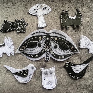 May include: A selection of handmade felt decorations, featuring a moth, bats, birds, a mushroom, and a shooting star. The ornaments are mainly black and white with embroidered details and small embellishments.