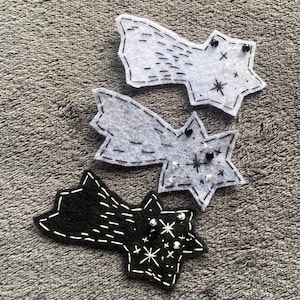 May include: Three felt star-shaped comet ornaments in white, gray, and black. Each has a star shape with a tail, detailed with white stitching. The gray and black stars have small bead accents, creating a celestial theme.