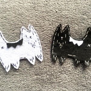 May include: Two cat-shaped felt ornaments. One is white with a black bow tie, the other black with a white bow tie. Both have white stitched details and an 'X' on the side.