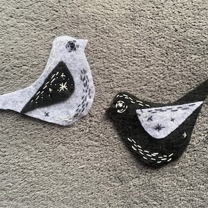 Puede incluir: Dos adornos de pájaros de fieltro. Uno es gris con un ala negra y el otro es negro con un ala gris. Ambos tienen detalles cosidos en blanco y acentos de estrellas. Los pájaros están hechos a mano.