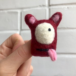 Könnte beinhalten: Eine kleine, rote Filz-Fingerpuppe mit einem weißen Gesicht, schwarzen Augen und einer herausgestreckten rosa Zunge. Die Puppe hat rosa Ohren und wird von einer Hand gehalten.