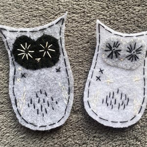 Puede incluir: Dos adornos de fieltro hechos a mano con forma de búho. Un búho tiene ojos negros con detalles blancos, el otro tiene ojos grises con detalles negros. Ambos tienen contornos cosidos en negro y costuras decorativas.