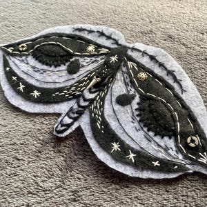 May include: A grey and black felt moth-shaped brooch with detailed embroidery. The design includes layered felt, white stitching, and decorative embellishments. The brooch is approximately 10 cm wide.