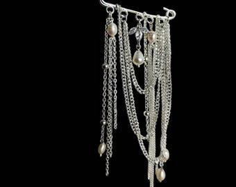 Puede incluir: Un broche de imperdible plateado con múltiples cadenas colgantes y detalles de perlas. El broche presenta un elemento floral decorativo y varias longitudes de cadenas delicadas, creando un efecto en capas. El fondo es negro sólido.