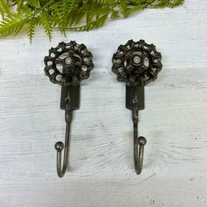 Może przedstawiać: Dwa haczyki ścienne w stylu antycznym z ciemnym brązowym wykończeniem. Każdy haczyk posiada dekoracyjną, okrągłą górę przypominającą koło zaworu i zakrzywiony haczyk. Haczyki są zamontowane na białej drewnianej powierzchni z zieloną paprocią.