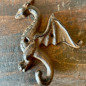 Może przedstawiać: Hak w kształcie smoka wykonany z żeliwa z brązowym wykończeniem. Smok znajduje się w pozycji lotu z rozpostartymi skrzydłami. Hak ma kształt ogona smoka.