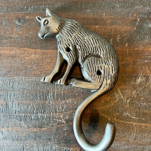 Może przedstawiać: Srebrny metalowy hak w kształcie kota ze zwiniętym ogonem. Hak został zaprojektowany tak, aby wyglądał jak kot siedzący na swoich zadach.