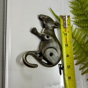 Może przedstawiać: Brązowa metalowa figurka szczura, o długości około 7.6 cm, ze zwiniętym ogonem i dwoma otworami na śruby. Żółta miarka znajduje się obok szczura, a w tle zielone liście przypominające paproć.