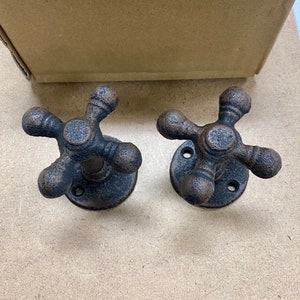 May include: Two antique-style faucet handles with a dark bronze finish. Each handle has a four-arm design and a circular base with mounting holes. The handles are made of metal and have a rustic, weathered appearance.