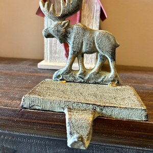 Peut inclure: Un porte-bas de Noël en fonte avec une figurine de wapiti détaillée dans une finition antique vieillie. L'élan se dresse sur une base rectangulaire avec un crochet pour suspendre les bas. Une petite maison décorative est en arrière-plan.