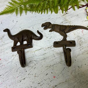 Może przedstawiać: Dwa haczyki w kształcie dinozaurów z żeliwa. Jeden haczyk ma kształt Tyrannosaurus Rex, a drugi Brontozaura. Oba haczyki są brązowe i mają rustykalne wykończenie.
