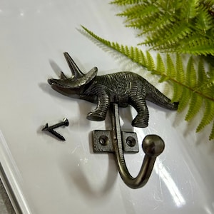 May include: A bronze-coloured metal wall hook shaped like a triceratops dinosaur. The hook has a curved design with a ball at the end. Two black screws are included for mounting. The hook is a decorative item.