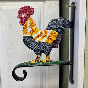 Może przedstawiać: Czarny metalowy uchwyt ścienny w kształcie koguta z zieloną podstawą i żółtymi i czerwonymi akcentami. Kogut jest zwrócony w prawo i ma czarno-białe wzorzyste ciało.