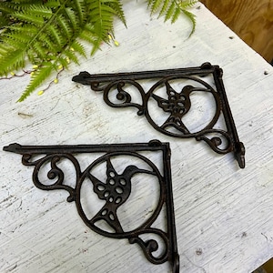 May include: Two dark brown cast iron shelf brackets. Each bracket features a hummingbird design within a circular frame, with decorative scrollwork. The brackets are designed to be mounted on a wall and support a shelf. The brackets are made of cast iron.